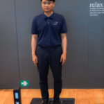 3D posture analysis for employees 2 - The Balance Studio - T.092 2469 462 Man in a navy polo shirt stands upright on a black measurement mat with markings in a room with gray-blue walls.