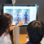 3D posture analysis for employees 3 - The Balance Studio - T.092 2469 462 Person and another figure view a large touchscreen displaying a posture analysis with blue diagrams and labeled body views; one person points at the screen.