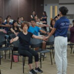 3D posture analysis for employees 4 - The Balance Studio - T.092 2469 462 Instructor in blue shirt leads a seated exercise class, raising participants' arms in front of them in a conference room setting with rows of chairs and an attentive group of adults.