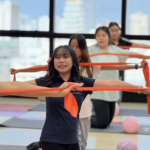 office syndrome workshop for employees 22 - The Balance Studio - T.092 2469 462 Group of women in a fitness class using red resistance bands in a bright studio by large windows overlooking a cityscape.